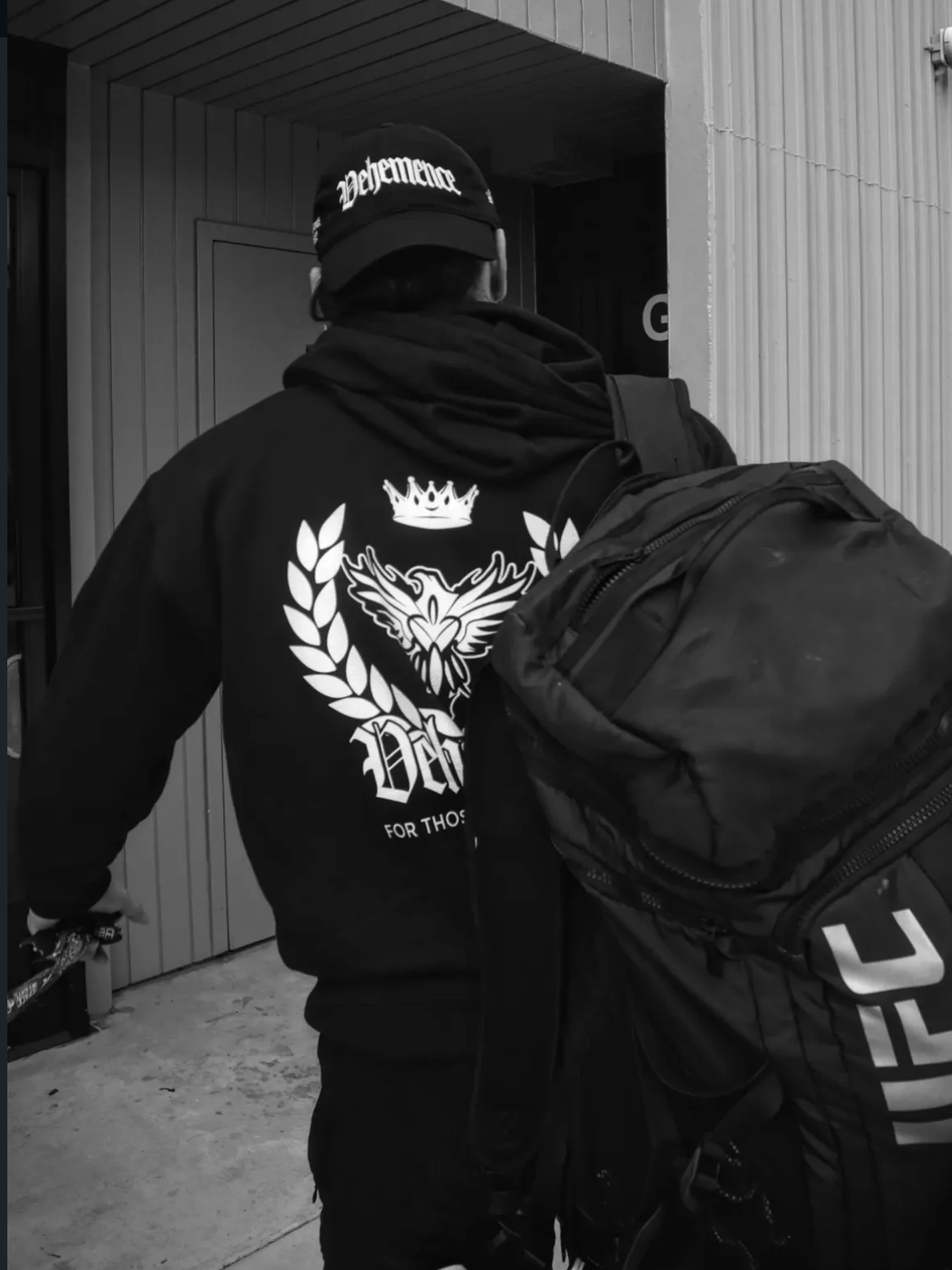 Person wearing a hoodie with a logo and UFC backpack, standing in front of a door.